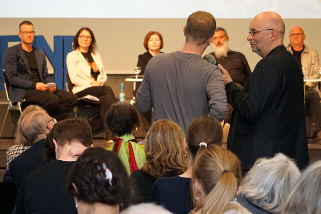 Zwei Männer stehen im Publikum. Der rechte hälte dem linken das Mikro hin. Man sieht beide von hinten. Auf der Bühne sind die Podiumsteilnehmer:innen unscharf zu sehen.