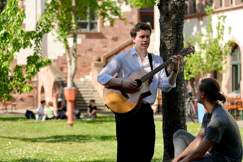 Ein Student steht mit einer Gitarre an einen Baum gelehnt, neben ihm sitzt ein anderer Student.