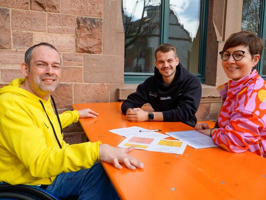 Das Foto zeigt Thorsten Lihl, Jan-Erik Möller und Sarah Maier im Gespräch. Sie sitzen an einem orange-farbenen Tisch im Außenbereich der Mensa der PH Heidelberg.