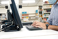 Auf dem Symbolbild sieht man links einen PC-Bildschirm und rechts die Hände eines Mannes, der auf der Tastatur tippt. Das Bild wurde im Lesesaal der Hochschule aufgenommen.