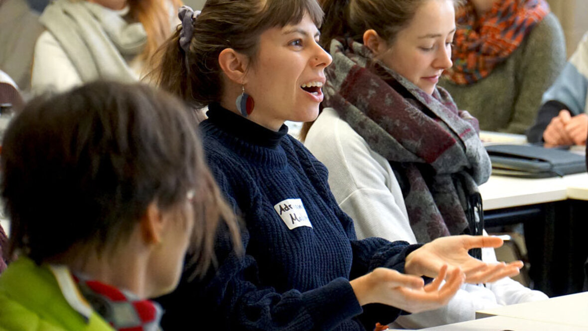 Eine junge Frau sitzt im vollen Seminarraum und spricht deutlich mit den Händen.