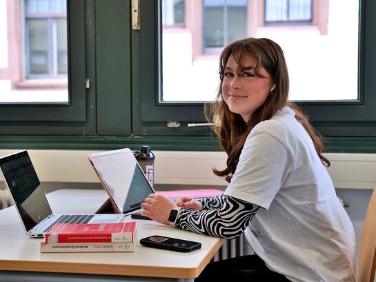 Zu sehen ist eine Studentin in der Bibliothek, die arbeitet und in die Kamera schaut