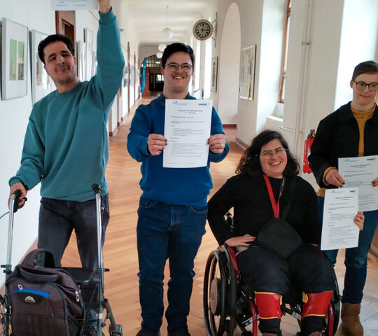 Das Bild zeigt die Qualifizierungsteilnehmenden Cedric Rüter, Lilly Lorenz, Maya Silva und Mehmet Can Friedel nach ihrer erfolgreichen Modulprfung auf einem Flur im Altbau der PH Heidelberg. Alle halten ein Zertifikat zur bestandenen Prüfung in die Luft.