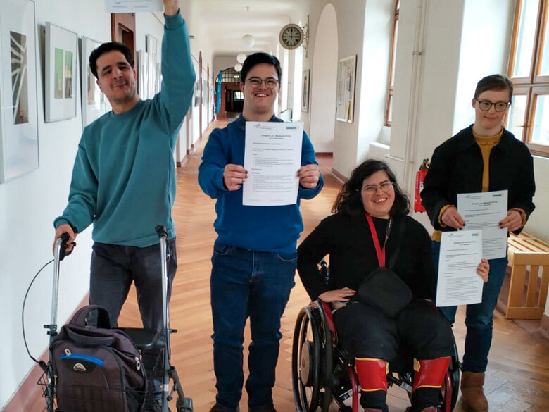 Das Bild zeigt die Qualifizierungsteilnehmenden Cedric Rüter, Lilly Lorenz, Maya Silva und Mehmet Can Friedel nach ihrer erfolgreichen Modulprfung auf einem Flur im Altbau der PH Heidelberg. Alle halten ein Zertifikat zur bestandenen Prüfung in die Luft.