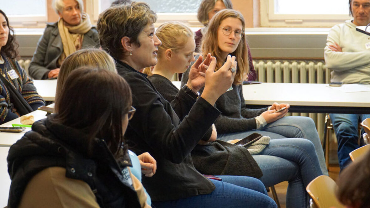 Eine Frau sitzt im vollen Seminarraum und spricht deutlich mit den Händen.