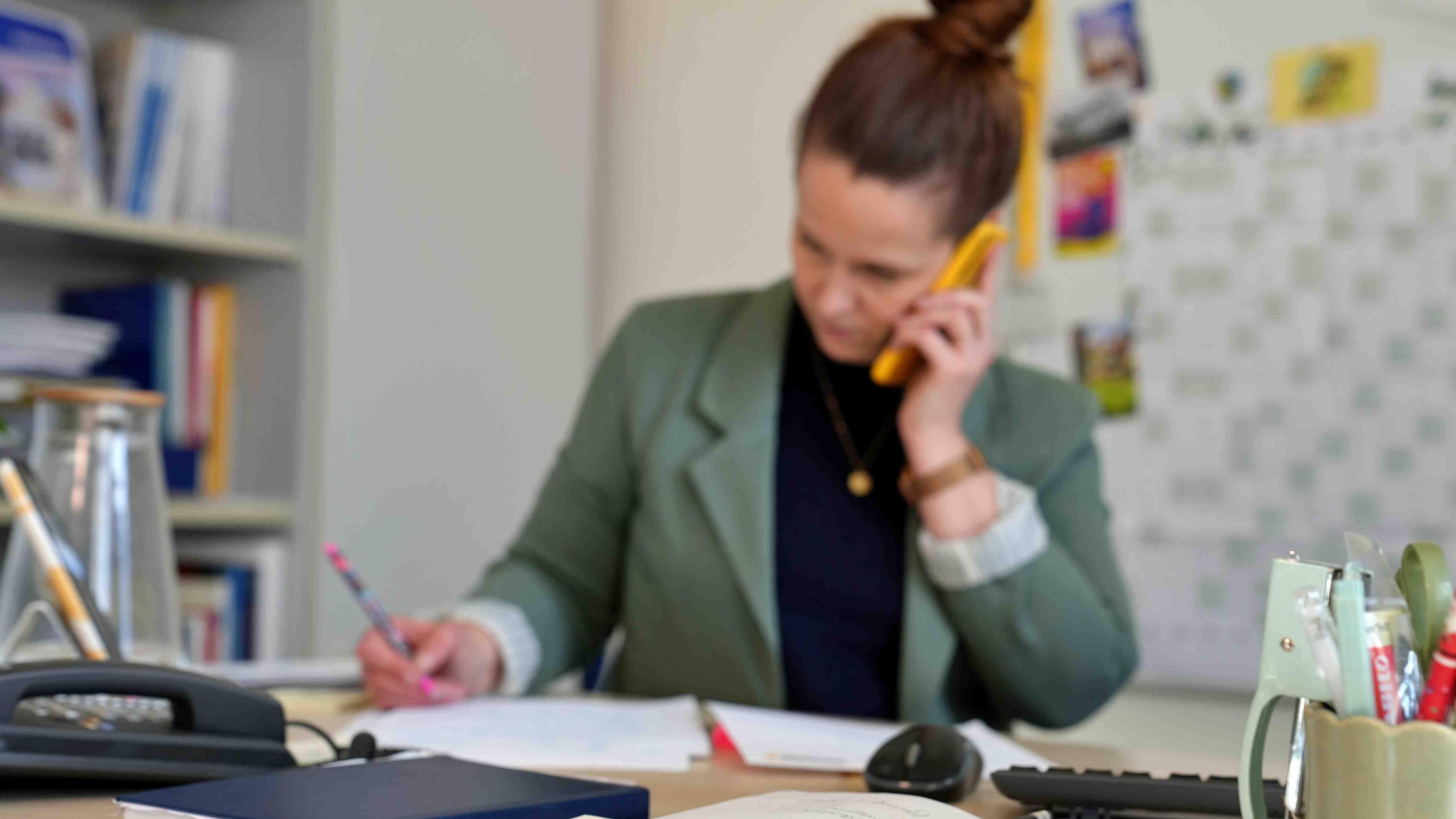 Eine Frau steht am Sitz-Steh-Tisch. Sie telefoniert mit dem Handy und notiert sich etwas. Der Fokus des Bildes liegt auf dem Schreibtisch, auf dem u.a. ein Festnetz und ein Kalender zu sehen sind.