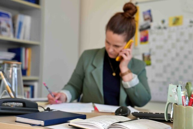 Eine Frau steht am Sitz-Steh-Tisch. Sie telefoniert mit dem Handy und notiert sich etwas. Der Fokus des Bildes liegt auf dem Schreibtisch, auf dem u.a. ein Festnetz und ein Kalender zu sehen sind.