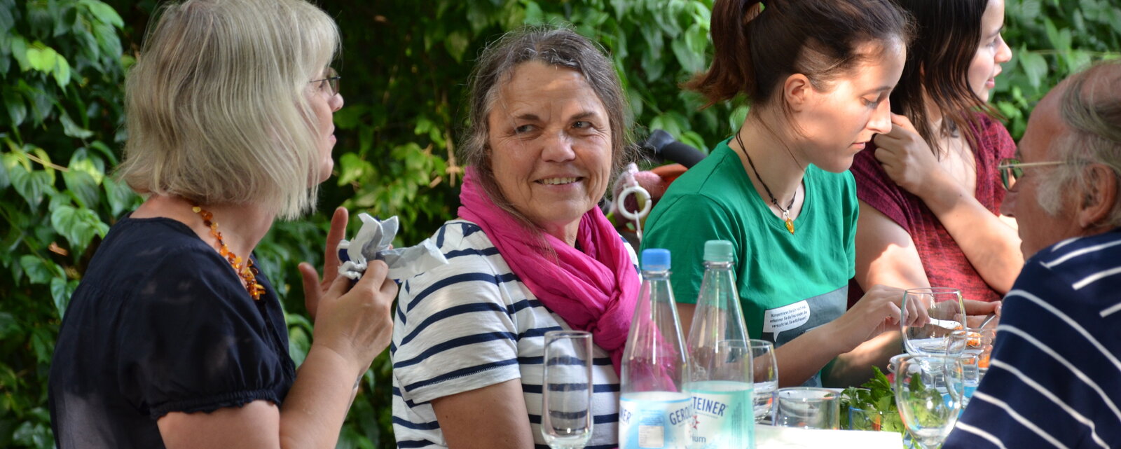 Mehrere Menschen sitzen im Ökogarten an einem Tisch und unterhalten sich.