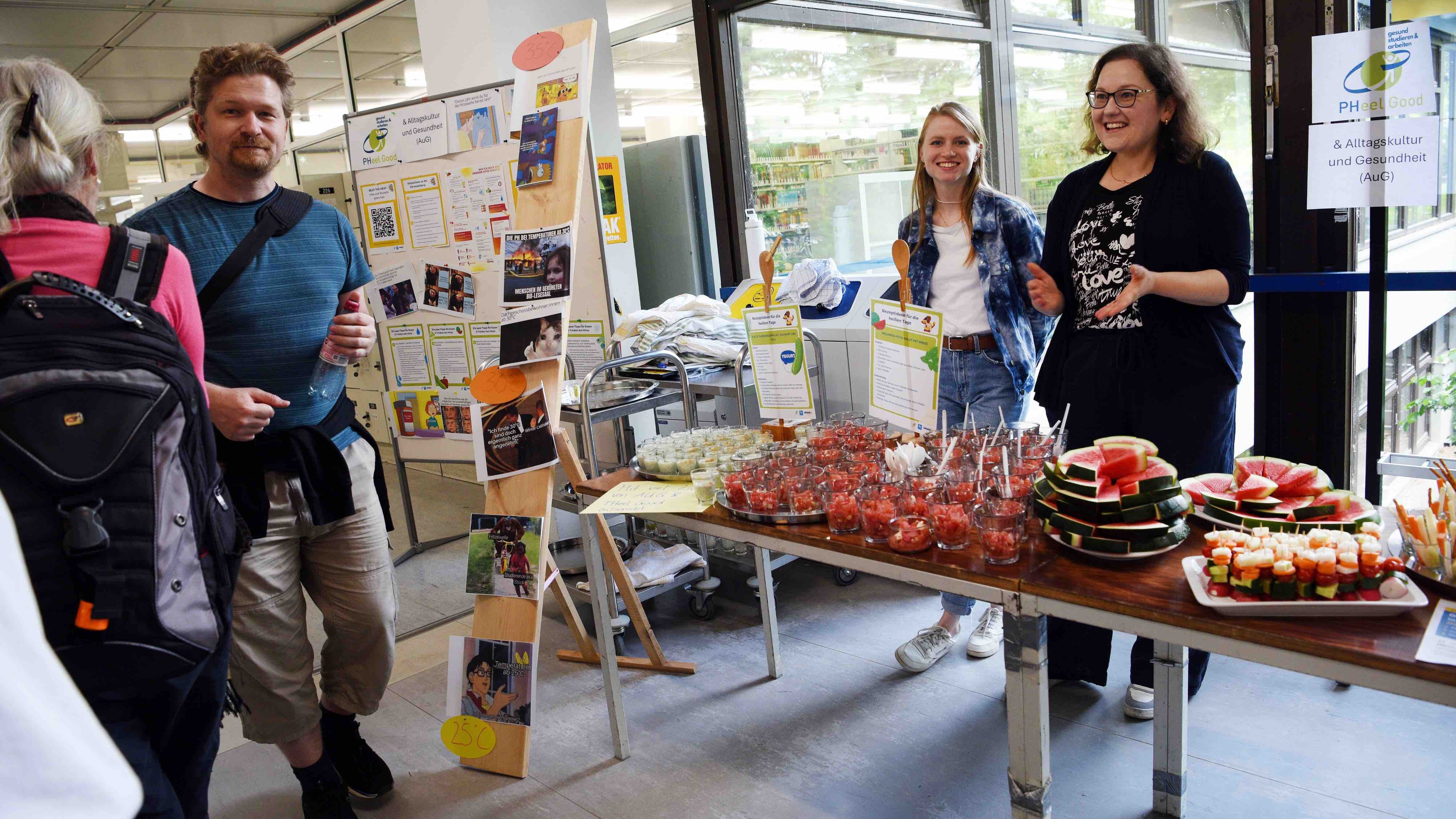 Das Team des Gesundheitsmanagement steht lachend hinter einem Tisch, auf dem viel gesundes Essen steht. Vor dem Tisch laufen mehrere Mitarbeiter:innen vorbei. Das Bild wurde im Neubaufoyer aufgenommen.