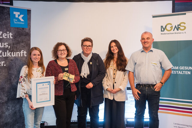 Fünf Menschen sind abgebildet. Links im Bild ist Hannah, sie hält eine gerahmte Urkunde in der Hand. Die Urkunde ist eine Auszeichnung für Partizipation im Gesundheitsmanagement. Rechts im Bild sind Jurymitglieder von der TK und der LVG & AfS