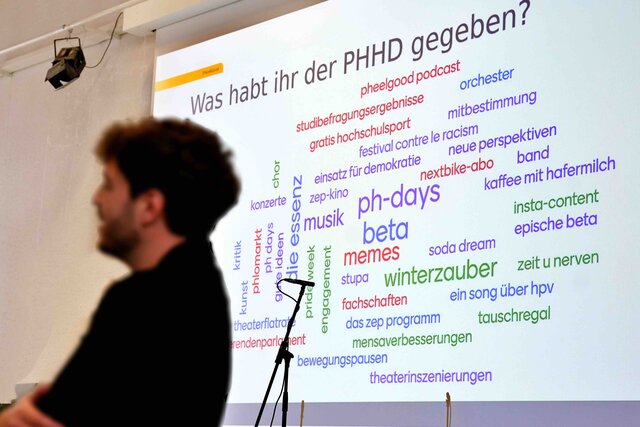 Zu sehen ist Max Wetterauer (etwas verschwommen & am Rednerpult stehend). Im Hintergrund ist eine Präsentationsseite projziert auf eine Leinwand zu sehen.