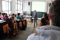 Lehrenden der sich an seine im Hörsaal sitzende Studierenden wendet.