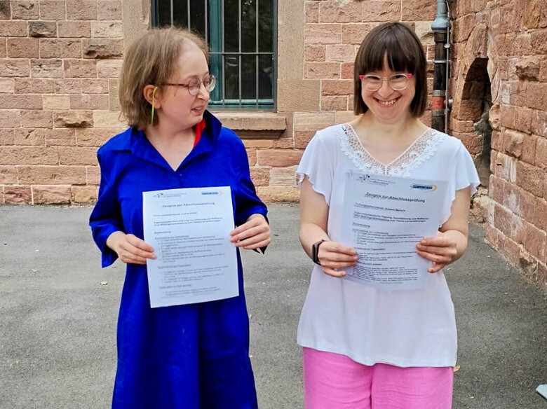 Louisa Kabbe und Susann Bensch stehen nebeneinander im Innenhof der PH Heidelberg. Sie halten ihre Zertifikate zur Abschlussprüfung in den Händen und lächeln in die Kamera.