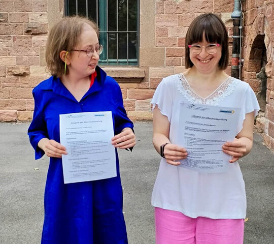 Louisa Kabbe und Susann Bensch stehen nebeneinander im Innenhof der PH Heidelberg. Sie halten ihre Zertifikate zur Abschlussprüfung in den Händen und lächeln in die Kamera.