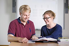 Das Foto zeigt zwei lachende Studierende die gemeinsam ein Buch lesen.