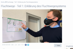 Das Bild zeigt einen Mann mit Maske der etwas an einem Plakat zeigt.