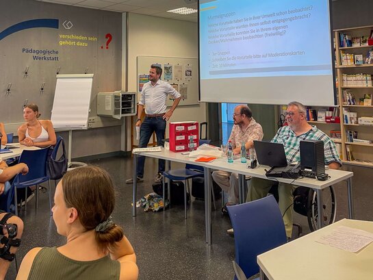Aufnahme der Seminarsitzung: Die Bildungsfachkräfte Thilo Krahnke und Helmuth Pflantzer sitzen an einem Tisch, Professor Böttinger steht neben ihnen. Sie sprechen zu Studierenden, die den dreien gegenüber sitzen.