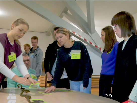 Bildungsfachkraft Susann Bensch steht gemeinsam mit Studierendem vor einem Tisch, auf dem Karten und ein Zeitstrahl liegen. 