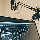 Auf dem Bild sieht man einen PC-Bildschirm mit Audio-Software und im Hintergrund ein Mikrofon im Podcaststudio.