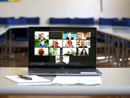 Aufnahme eines Laptops, der auf einem Tisch im Senatssaal der PH Heidelberg steht. Auf dem Bildschirm des Laptops sind viele kleine Bilder einer Video-Konferenz.