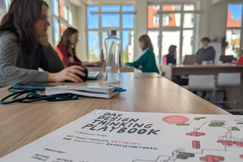 Zu sehen ist ein Buch über Design Thinking, dahinter Menschen am Arbeiten.