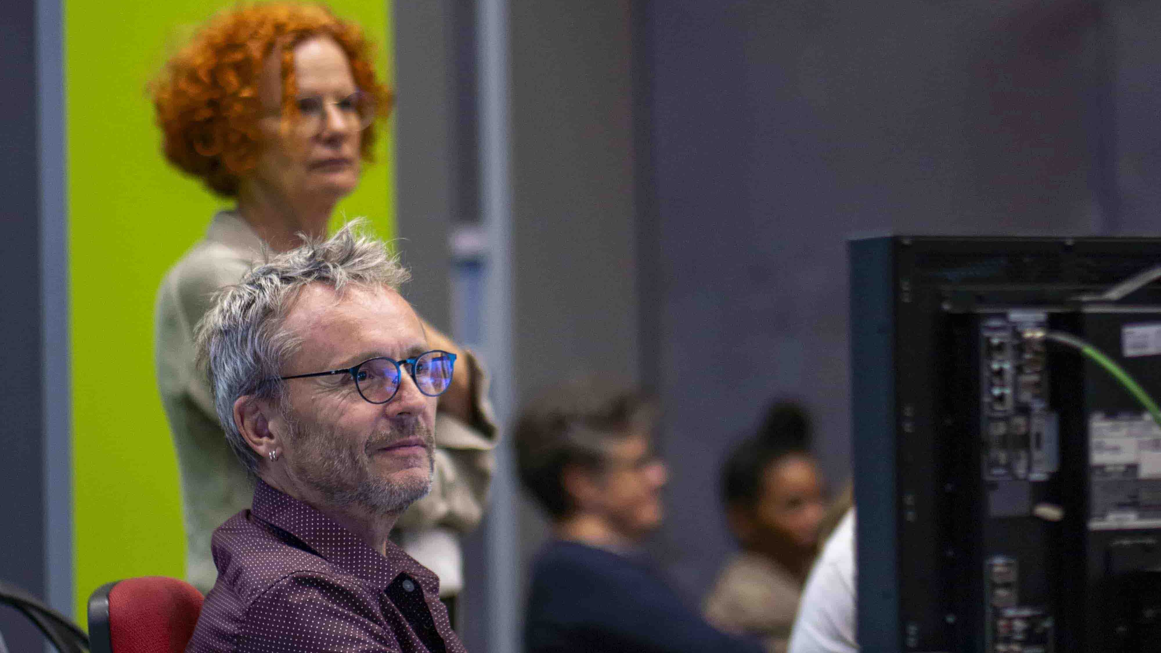 Zwei Personen sind vor einem Laptop und Monitoren.