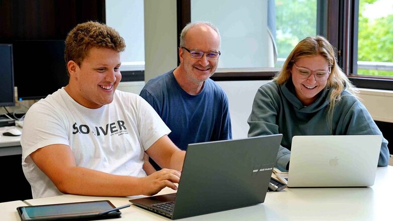 Zwei Studierende und ein Dozent der Mathematik sitzen lächelnd an einem Tisch. Vor ihnen stehen zwei Laptops.