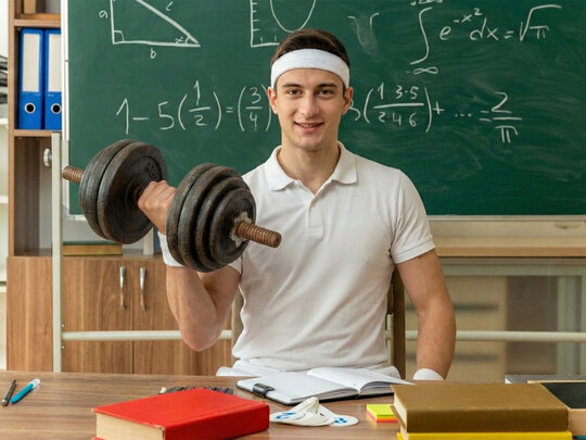 Das Bild zeigt einen fitten jungen Lehrer mit Hantel in der Hand vor einer Tafel am Lehrerpult.
