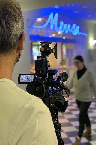 Das Foto zeigt im Vordergrund einen Mann mit einer Videokamera. Im Hintergrund läuft eine Bildungsfachkraft.