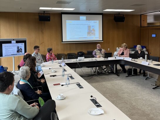 Aufnahme vom sozialpolitischen Frühstück. Mehrere Personen sitzen im Halbkreis an Tischen, Bildungsfachkraft Helmuth Pflantzer spricht und im Hintergrund ist eine PowerPoint-Präsentation, auf deren Seiten steht "Ziele meiner Arbeit als Bildungsfachkraft".