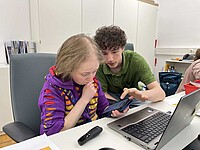 Schülerpraktikant Sander Vach sitzt mit Qualfizierungstelnehmerin Louisa Kabbe am Schreibtisch. Sie hält ein Smartphone in der Hand, er erklärt ihr etwas zur Funktionsweise.