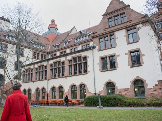 Eine Frau im roten Mantel blickt auf den Altbau. Sie ist lediglich von hinten zu sehen. Das Bild wurde im Innenhof im Winter aufgenommen.