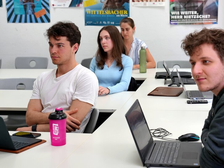 Studierende des Fachs Geschichte auf Lehramt sitzen in einem Seminar und hören dem Dozenten zu. Auf den Tischen stehen Laptops und Schreibmaterialien.