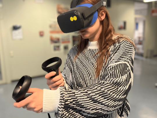 [Übersetzen nach: English] Eine junge Frau der Pädagogischen Hochschule Heidelberg nutzt eine VR-Brille und Controller, um in eine digitale Umgebung einzutauchen.