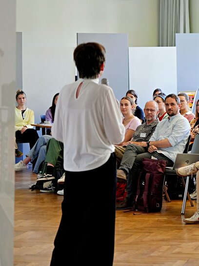 Zu Sehen ist eine Frau die vor Publikum eine Rede hält. 