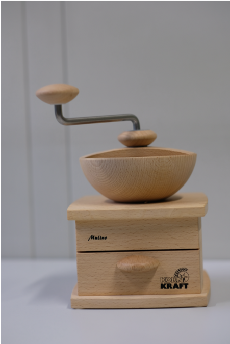 Die Getreidemühle aus Holz ähnelt vom Aussehen einer Kaffeemühle. Gibt man in die obere Öffnung Körner hinein und dreht anschließend an der Kurbel, wird daraus feines Mehl gemahlen. Eine genauere Beschreibung liefert der Text unterhalb des Bildes.