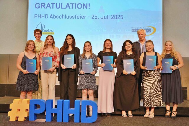 Zu sehen sind Alumni, die auf einer Bühne stehen und ihre Bestehensbriefe in PHHD-Mappen hochhalten. Gemeinsam mit Rektorin Vach & Prorektor Dammer (a.D.), welche ihnen die Mappen überreicht haben.