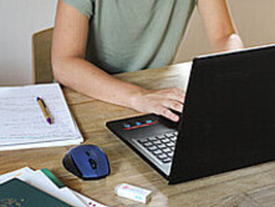 Das Bild zeigt eine Person, die an einem Tisch sitzt und mit einem Laptop arbeitet. Auf dem Tisch liegen auch noch ein Block und weitere Schreibutensilien.
