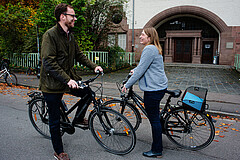 Auf dem Bild sieht man einen Mann und eine Frau. Beide sitzen auf ihre stehenden Fahrräder und unterhalten sich. Das Foto wurde vor dem Altbau der Hochschule aufgenommen. 