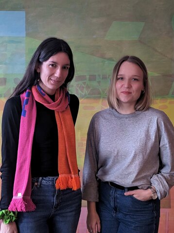 Zu sehen sind Alina von Heyn & Marleen Langer vor einer bunten Wand.