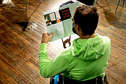 Das Foto zeigt eine Person im Rollstuhl von hinten oben. Diese Person liest gerade ein Magazin.