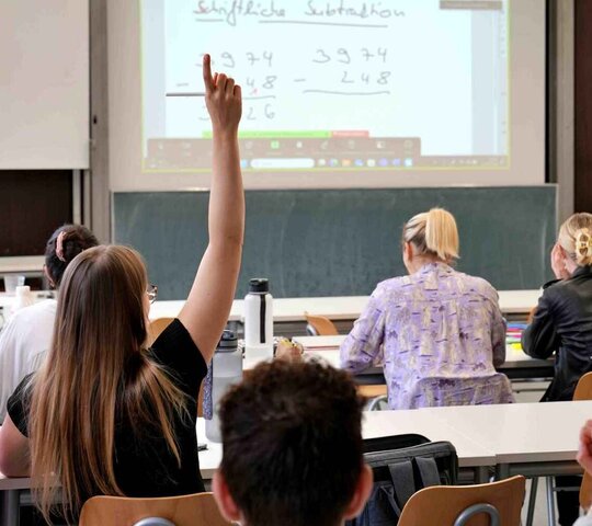 Zu sehen ist eine Gruppe Studierender in einem Seminarraum. Eine Person meldet sich.