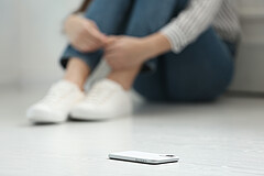 Das Symbolbild zeigt vorne ein Handy auf dem Boden. Im Hintergrund sieht man unscharf eine Person sitzen, die die Arme eng um ihre Beine schlingt. Copyright: Adobe Stock.