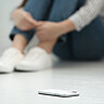 Das Symbolbild zeigt vorne ein Handy auf dem Boden. Im Hintergrund sieht man unscharf eine Person sitzen, die die Arme eng um ihre Beine schlingt. Copyright: Adobe Stock.