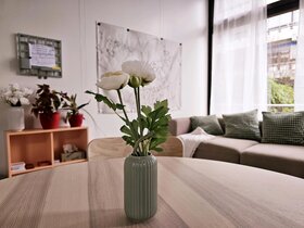 Abgebildet ist der Personalraum. Im Vordergrund steht eine Blumevase mit einer Blume auf dem Esstisch, im Hintergrund ist ein Sofa mit Kossen. Dahinter ist eine große Fensterfront mit Blick ins Grüne