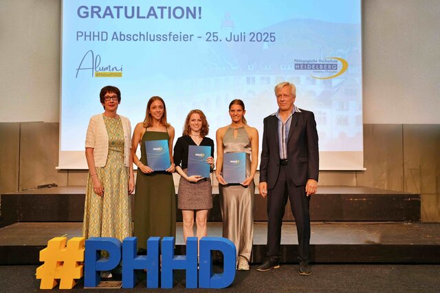 Zu sehen sind Alumni, die auf einer Bühne stehen und ihre Bestehensbriefe in PHHD-Mappen hochhalten. Gemeinsam mit Rektorin Vach & Prorektor Dammer (a.D.), welche ihnen die Mappen überreicht haben.
