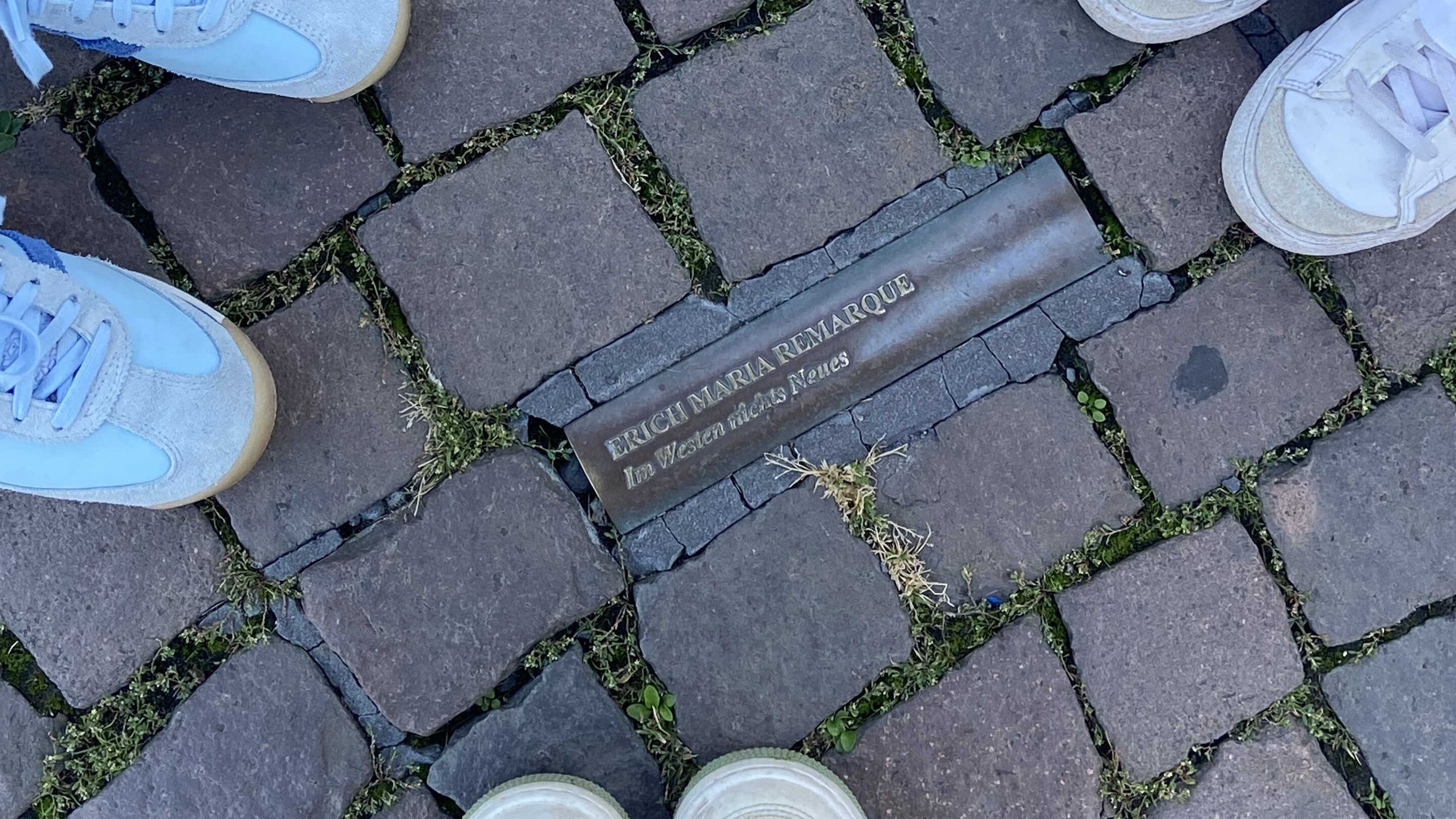 Foto eines Namenschildes in den Plastersteinen und Füßen drum herum