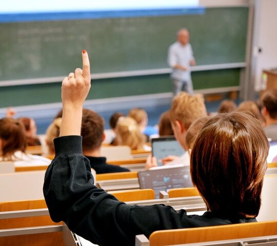 Zu sehen ist ein Hörsaal mit Studierenden, eine von ihnen meldet sich mit der Hand
