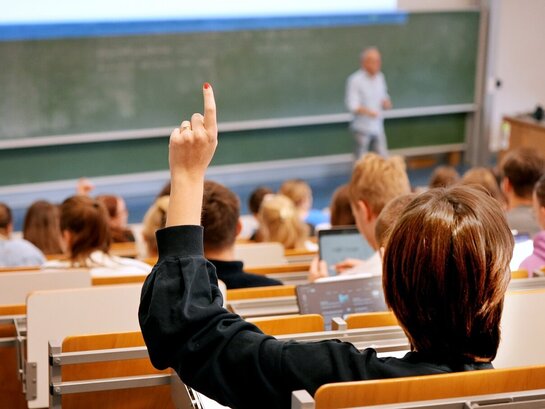Zu sehen ist ein Hörsaal mit Studierenden, eine von ihnen meldet sich mit der Hand