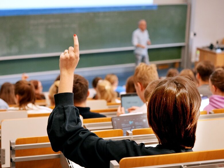 Zu sehen ist ein Hörsaal mit Studierenden, eine von ihnen meldet sich mit der Hand